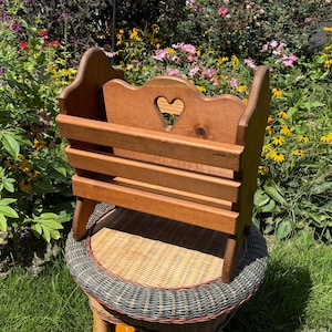May include: A medium-brown wooden magazine rack featuring a slatted front design and a solid back panel with a heart-shaped cutout at the top center. The rack has curved side supports and a sturdy base, constructed from natural wood with a smooth finish.