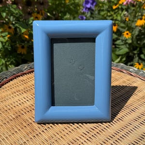 May include: A rectangular picture frame with a smooth, glossy light blue finish. The frame features a thick, beveled border surrounding a dark gray insert. The design is simple and uniform, with clean edges and a reflective surface on the frame.