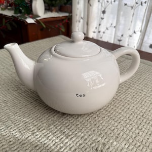 May include: A white ceramic teapot with a lid and handle. The word "tea" is printed in black on the front of the teapot. The teapot is on a textured, light-coloured surface.