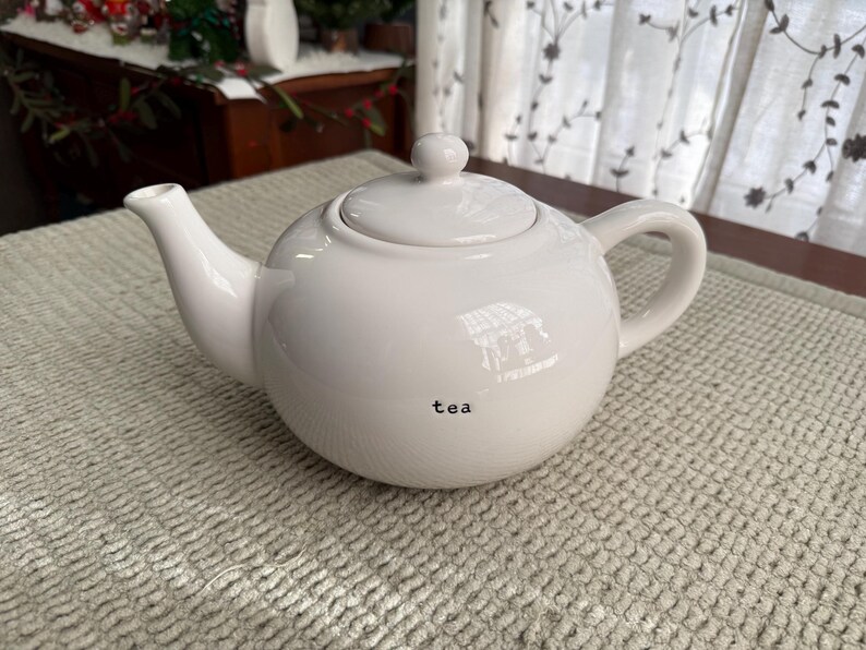 May include: A white ceramic teapot with a lid and handle. The word "tea" is printed in black on the front of the teapot. The teapot is on a textured, light-coloured surface.