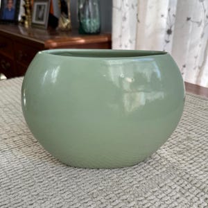 May include: A round, sage green ceramic bowl with a glossy finish. The bowl has a wide opening and a rounded shape, suitable for decorative or functional use. The bowl is sitting on a textured, light-colored surface.