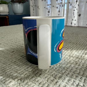 Peut inclure: Une tasse en c&eacute;ramique blanche avec une anse noire et le texte "But First, Coffee" en gras noir. La tasse est pos&eacute;e sur un fond flou, sugg&eacute;rant un cadre chaleureux.