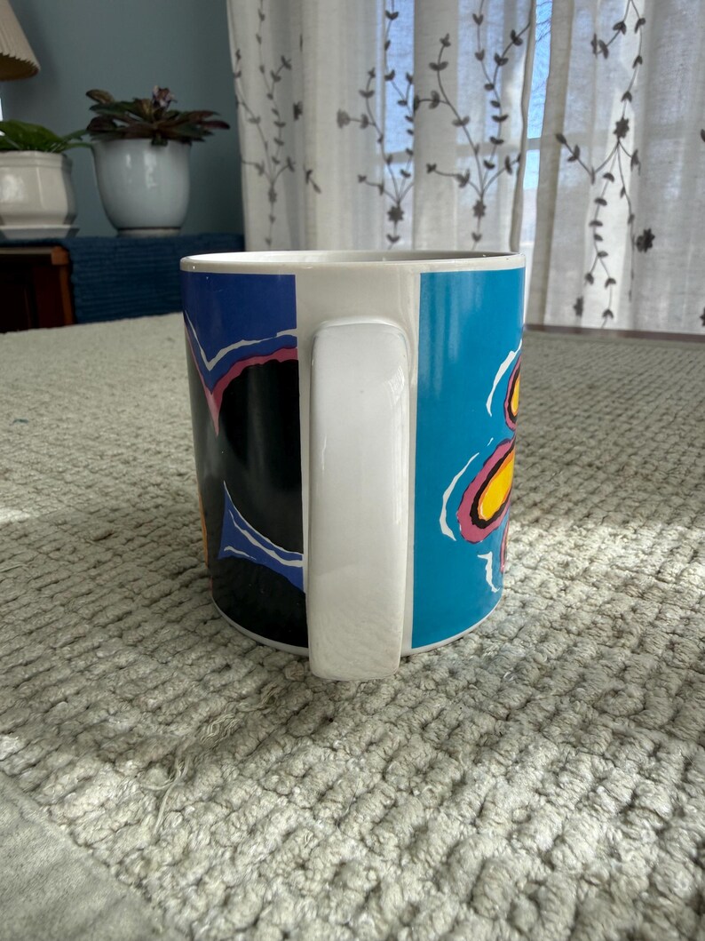 Peut inclure: Une tasse en c&eacute;ramique blanche avec une anse noire et le texte "But First, Coffee" en gras noir. La tasse est pos&eacute;e sur un fond flou, sugg&eacute;rant un cadre chaleureux.