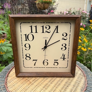 May include: A square wall clock with a dark brown wooden frame and a cream-colored face. The clock features black Arabic numerals from 1 to 12, black hour and minute hands, and the Sunbeam logo printed in black text above the center hands.