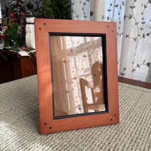 May include: A rectangular wooden picture frame with a reddish-brown finish. The frame has a black interior border and small, dark-colored accents at each corner. The frame is resting on a textured, light-colored surface.