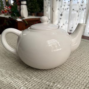 May include: A white ceramic teapot with a rounded body, a curved handle, and a spout. The teapot has a lid with a small knob on top. The teapot is on a textured, light-coloured surface.