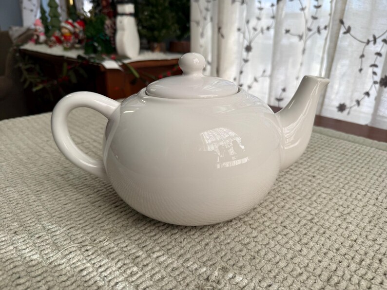 May include: A white ceramic teapot with a rounded body, a curved handle, and a spout. The teapot has a lid with a small knob on top. The teapot is on a textured, light-coloured surface.