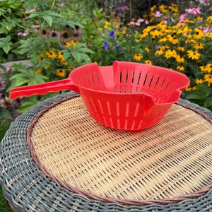 May include: A bright red plastic colander featuring a long handle with a textured grip, a deep bowl shape, and vertical drainage slots along the sides. The kitchen tool has a notched rim design and is constructed from a single piece of molded material.