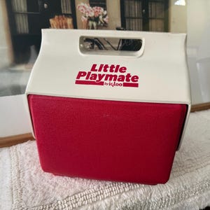 May include: A vintage Little Playmate by Igloo cooler. The cooler has a white top with a handle and a red base. The words "Little Playmate" are printed in red on the front. The cooler is sitting on a white towel.