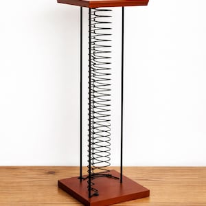 May include: A unique, modern CD or record holder. It features a dark brown wooden base and top, connected by black metal supports and a central spring design. The holder is designed to stand on a flat surface.