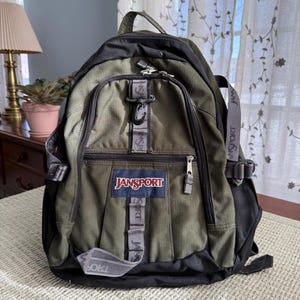 May include: Olive green and black Jansport backpack with multiple compartments and straps. The backpack features the Jansport logo in a blue rectangle with red lettering. The backpack has a top handle and adjustable shoulder straps.
