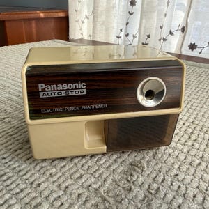 May include: A vintage Panasonic electric pencil sharpener. The sharpener has a beige plastic base and a dark wood-grain top with the text "Panasonic AUTO-STOP ELECTRIC PENCIL SHARPENER". The sharpener has a single sharpening hole.