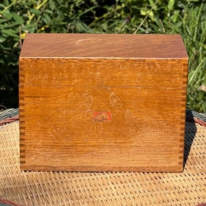 May include: A rectangular wooden box with a natural grain finish and visible finger joints at the corners. The front panel features a small, rectangular red label with indistinct black text centred on the surface of the wood.
