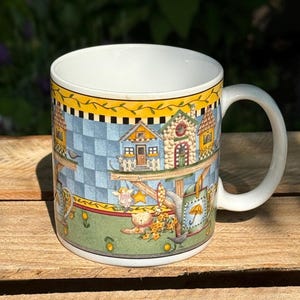 May include: White ceramic mug with a whimsical village scene. The mug features a yellow rim with a black checkered pattern, and a colorful illustration of houses, characters, and a cat. The mug has a white handle.