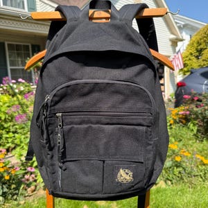 May include: A black backpack featuring a large main compartment, a smaller front zippered pocket, and a secondary front pocket. A circular logo with an eagle graphic and the text Eddie Bauer is visible on the lower right side of the front pocket.