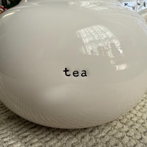 May include: A white ceramic teapot with a rounded body, a curved handle, and a spout. The word "tea" is printed in black on the front of the teapot. The teapot is sitting on a textured, light-coloured surface.