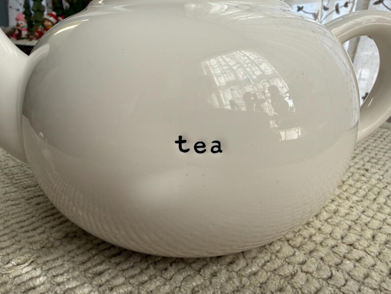 May include: A white ceramic teapot with a rounded body, a curved handle, and a spout. The word "tea" is printed in black on the front of the teapot. The teapot is sitting on a textured, light-coloured surface.