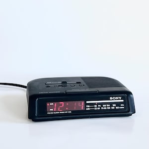 May include: A black Sony Dream Machine AM/FM clock radio with a red digital display showing the time as 12:14 PM. The radio has a speaker grill and control buttons on top.