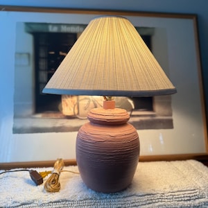 May include: A table lamp with a textured, dusty pink ceramic base and a conical, pleated fabric lampshade in a light beige tone. The lamp includes a brown electrical cord with a switch and a gold-toned plug, resting on a white surface.