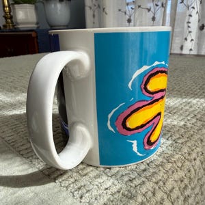 Peut inclure: Tasse en c&eacute;ramique blanche avec une grande anse incurv&eacute;e. La tasse pr&eacute;sente un motif vibrant avec un fond bleu et un motif floral jaune, rose et noir. La tasse est pos&eacute;e sur une surface textur&eacute;e gris clair.