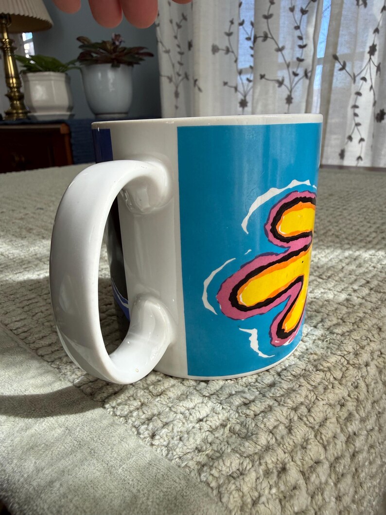 Peut inclure: Tasse en c&eacute;ramique blanche avec une grande anse incurv&eacute;e. La tasse pr&eacute;sente un motif vibrant avec un fond bleu et un motif floral jaune, rose et noir. La tasse est pos&eacute;e sur une surface textur&eacute;e gris clair.