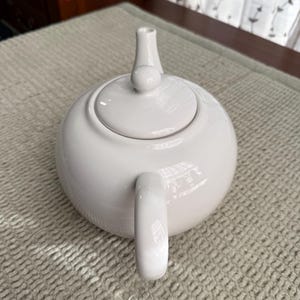 May include: A white ceramic teapot with a rounded body, a curved handle, and a lid with a small knob. The spout is slender and cylindrical. The teapot is on a textured, light-coloured surface.