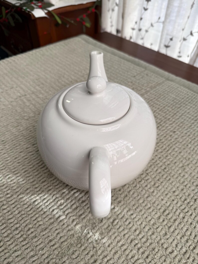 May include: A white ceramic teapot with a rounded body, a curved handle, and a lid with a small knob. The spout is slender and cylindrical. The teapot is on a textured, light-coloured surface.