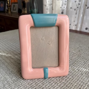 May include: A rectangular picture frame with a glossy pink finish and teal accents. The frame has a simple, modern design with a smooth surface and a neutral-colored backing for the photo.