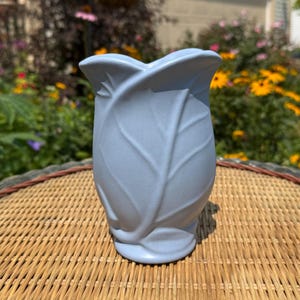 May include: A light blue ceramic vase with a matte finish, featuring a sculpted design that mimics the overlapping layers of a cabbage leaf. The vase has a wide, undulating rim and a tapered base, with raised veins detailing the leaf-like exterior.