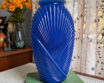 May include: A vibrant cobalt blue vase with a fan-like design. The vase is made of glass and has a glossy finish. It is sitting on a green book. The vase is empty and ready to be filled with flowers.