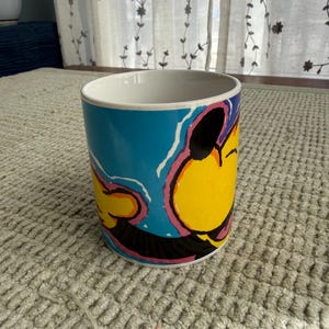 Peut inclure: Une tasse &agrave; caf&eacute; en c&eacute;ramique avec un dessin de personnage de dessin anim&eacute;. La tasse pr&eacute;sente un visage jaune avec des accents noirs, roses et bleus. L'int&eacute;rieur de la tasse est blanc et elle repose sur une surface textur&eacute;e de couleur claire.