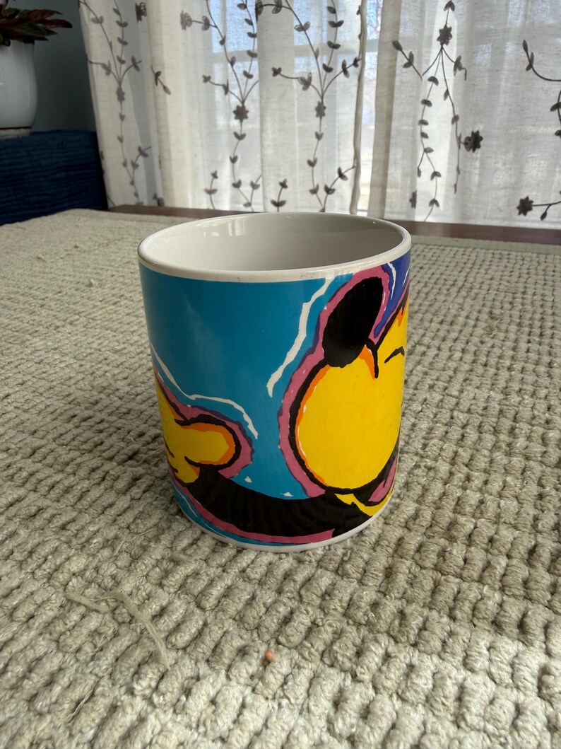 Peut inclure: Une tasse &agrave; caf&eacute; en c&eacute;ramique avec un dessin de personnage de dessin anim&eacute;. La tasse pr&eacute;sente un visage jaune avec des accents noirs, roses et bleus. L'int&eacute;rieur de la tasse est blanc et elle repose sur une surface textur&eacute;e de couleur claire.