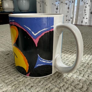 Peut inclure: Mug en c&eacute;ramique blanche avec un motif abstrait color&eacute;. Le mug pr&eacute;sente un fond noir avec des formes jaunes, oranges, bleues et roses. La poign&eacute;e est blanche et le mug repose sur une surface textur&eacute;e.