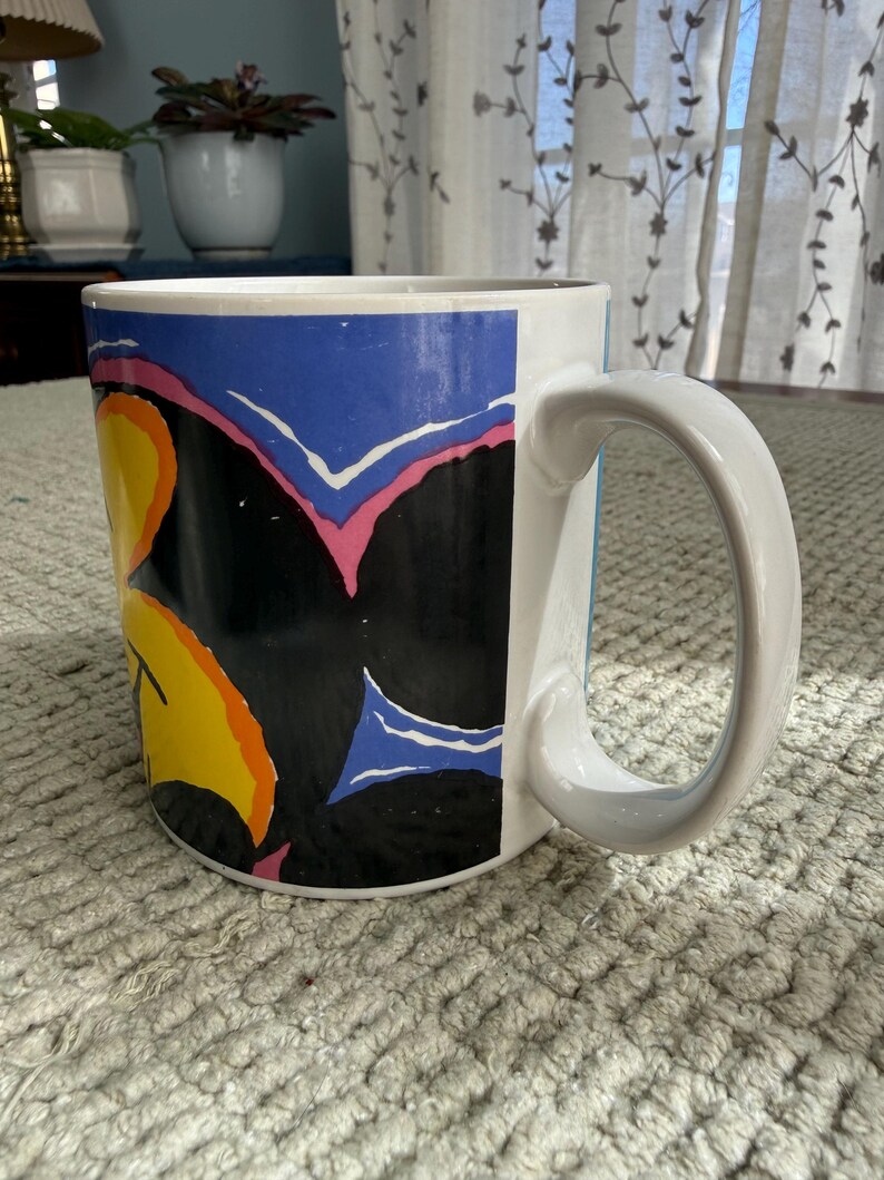 Peut inclure: Mug en c&eacute;ramique blanche avec un motif abstrait color&eacute;. Le mug pr&eacute;sente un fond noir avec des formes jaunes, oranges, bleues et roses. La poign&eacute;e est blanche et le mug repose sur une surface textur&eacute;e.