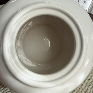May include: A white ceramic container with a wide, rounded rim and a deep, circular interior. The interior is a lighter shade of white, creating a contrast with the outer rim. The container has a smooth, glossy finish.