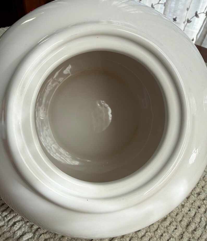 May include: A white ceramic container with a wide, rounded rim and a deep, circular interior. The interior is a lighter shade of white, creating a contrast with the outer rim. The container has a smooth, glossy finish.