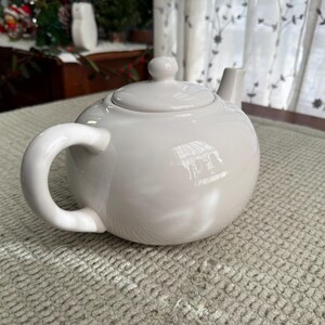 May include: A white ceramic teapot with a rounded body, a curved handle, and a small spout. The lid has a small knob. The teapot sits on a textured, light-coloured surface. The teapot is designed for brewing tea.