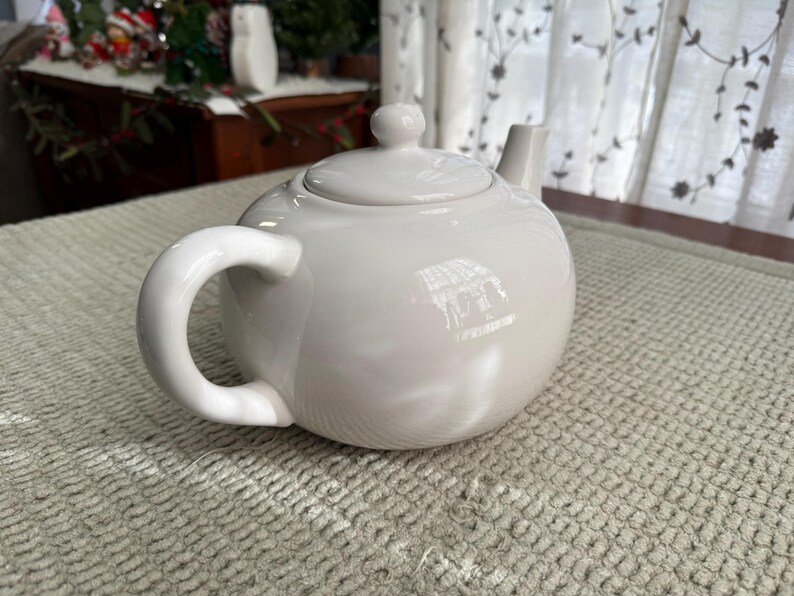 May include: A white ceramic teapot with a rounded body, a curved handle, and a small spout. The lid has a small knob. The teapot sits on a textured, light-coloured surface. The teapot is designed for brewing tea.