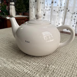 May include: A white ceramic teapot with a lid and handle, featuring the word "tea" in black lettering. The teapot is on a textured, light-coloured surface. The background includes a window with sheer curtains.