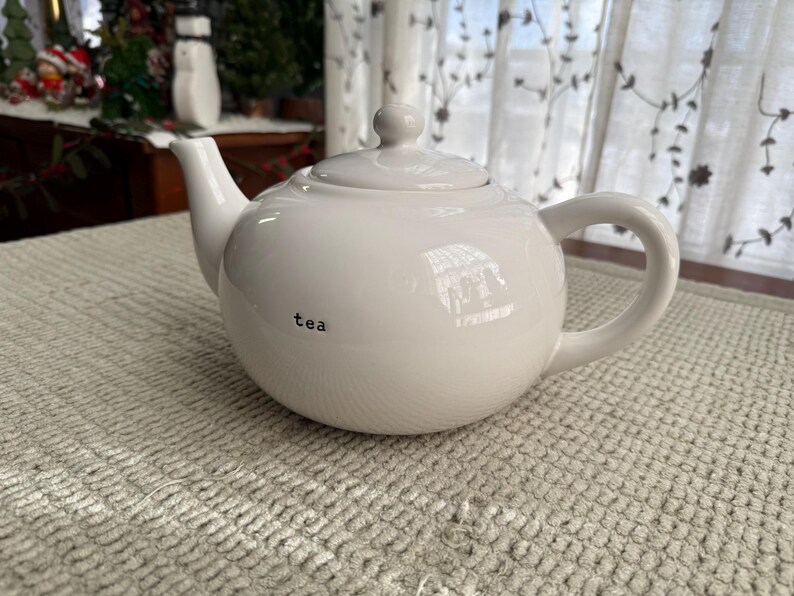 May include: A white ceramic teapot with a lid and handle, featuring the word "tea" in black lettering. The teapot is on a textured, light-coloured surface. The background includes a window with sheer curtains.
