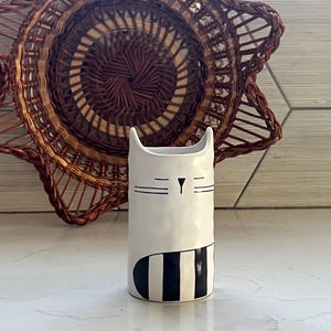 May include: A white ceramic vase with a cat face design. The vase has a cylindrical shape with a black and white striped pattern at the base. The background features a woven brown basket and a tiled wall.