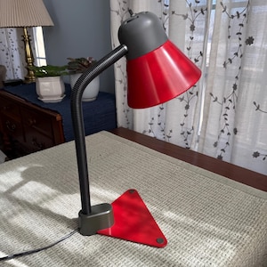 May include: A red and gray desk lamp with a flexible black neck and a triangular base. The lamp has a cone-shaped shade and is positioned on a light-colored surface.