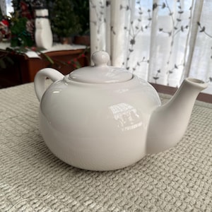 May include: A white ceramic teapot with a rounded body, a curved spout, and a loop handle. The lid has a small knob. The teapot sits on a textured, light-coloured surface. The background includes a window with sheer curtains.
