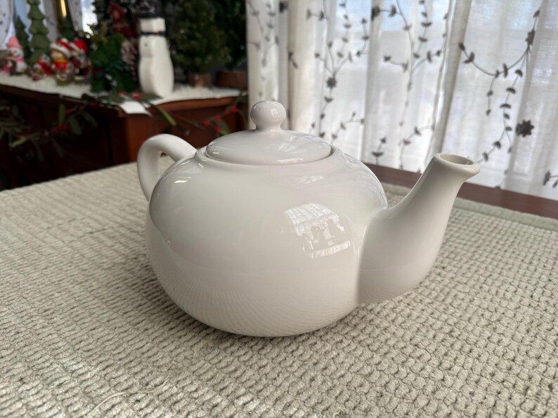 May include: A white ceramic teapot with a rounded body, a curved spout, and a loop handle. The lid has a small knob. The teapot sits on a textured, light-coloured surface. The background includes a window with sheer curtains.