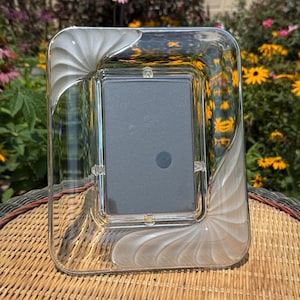 May include: A rectangular picture frame made of clear glass with white, swirling, shell-like patterns at the top-left and bottom-right corners. The frame features a central rectangular opening for a photograph, held by small metal clips.