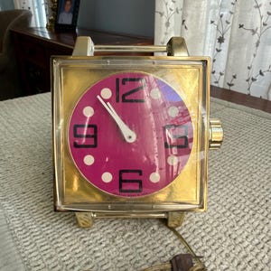 May include: A vintage gold-toned clock with a square face and a magenta clock face. The clock features large black numerals and white dots. The clock has a handle and a power cord. The clock is marked "Spartus Made in USA".