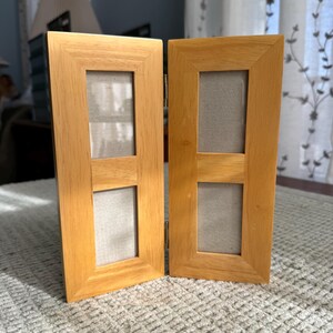 May include: Two light-coloured wooden photo frames, each with two rectangular openings. The frames are hinged together and stand upright on a textured surface. The frames have a natural wood grain finish.