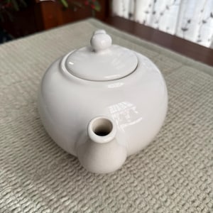 May include: A white ceramic teapot with a rounded body, a lid with a small knob, and a spout. The teapot is on a light-coloured textured surface. The teapot is designed for brewing tea.