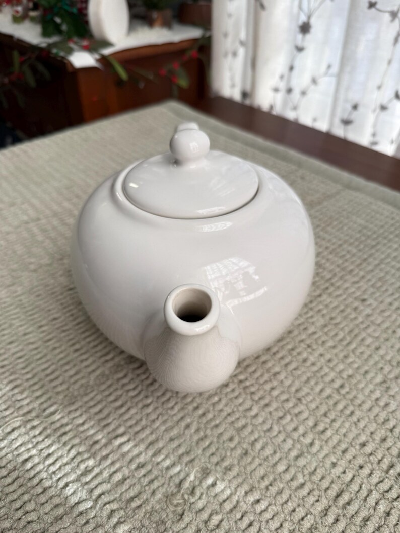 May include: A white ceramic teapot with a rounded body, a lid with a small knob, and a spout. The teapot is on a light-coloured textured surface. The teapot is designed for brewing tea.