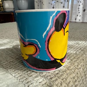 Peut inclure: Une tasse cylindrique en c&eacute;ramique avec un design pop art vibrant. La tasse pr&eacute;sente un fond bleu vif avec un motif de personnage de dessin anim&eacute; jaune, noir, rose et blanc. La tasse a un bord et une base blancs.